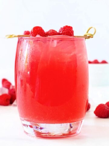 Close up front-angle hero shot of a glass filled to the brim with raspberry iced tea, garnished with a skewer holding three fresh raspberries. Scattered fresh raspberries and ice are behind the glass, with another iced raspberry tea filled glass and a white bowl of fresh raspberries blurred in the bright white background, all set on a white and gray marble countertop.