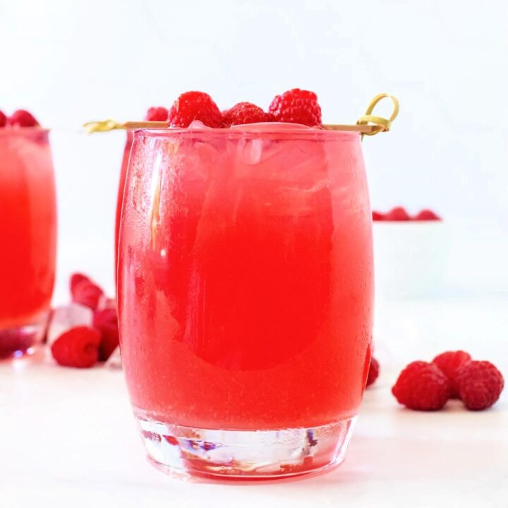 Close up front-angle hero shot of a glass filled to the brim with raspberry iced tea, garnished with a skewer holding three fresh raspberries. Scattered fresh raspberries and ice are behind the glass, with another iced raspberry tea filled glass and a white bowl of fresh raspberries blurred in the bright white background, all set on a white and gray marble countertop.