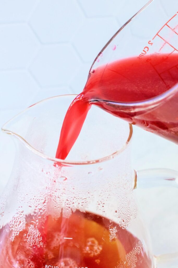 Glass measuring cup pouring hot raspberry juice mixture into a glass pitcher filled with steaming herbal tea.