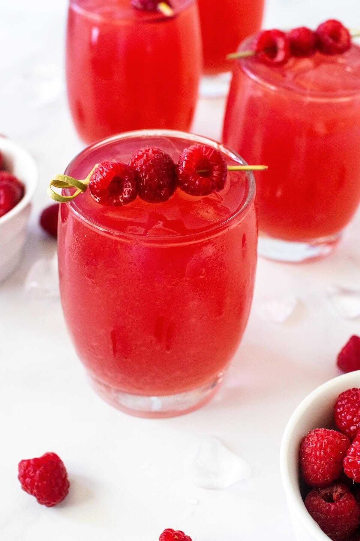 Glass filled to the brim with iced raspberry tea, garnished with a skewer holding three fresh raspberries. In the background, three more glasses of iced raspberry tea, each garnished with the same skewer, along with two white bowls filled with fresh raspberries, scattered raspberries, and ice cubes, all arranged on a white and gray marble countertop.