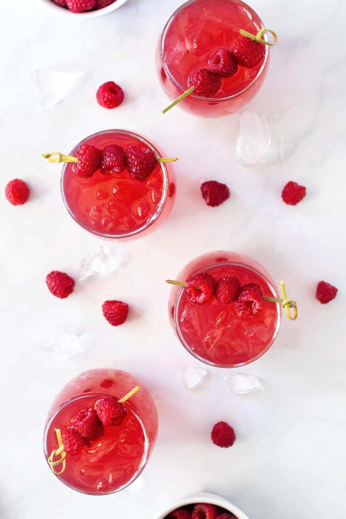 Overhead shot of four glasses filled to the brim with raspberry iced tea, each garnished with a skewer holding three fresh red raspberries. Fresh raspberries and ice cubes are scattered around the glasses on a white countertop surface, with two white bowls of raspberries partially visible off to the sides.