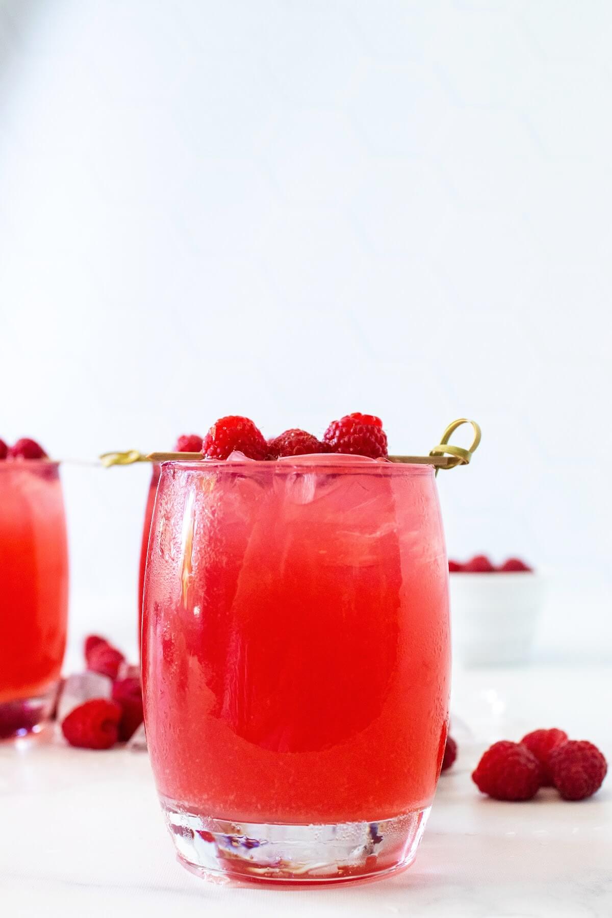 Front-angle hero shot of a glass filled to the brim with raspberry iced tea, garnished with a skewer holding three fresh raspberries. Scattered fresh raspberries and ice are behind the glass, with another iced raspberry tea filled glass and a white bowl of fresh raspberries blurred in the bright white background, all set on a white and gray marble countertop.