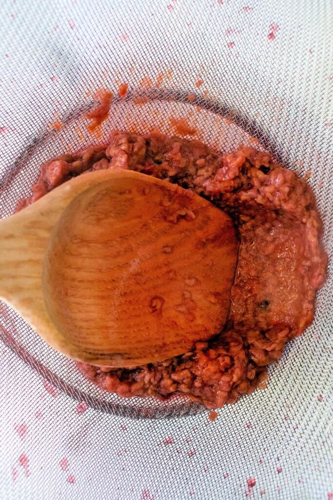 Overhead view of a large fine mesh strainer with a wooden spoon mashing cooked raspberry mixture over a glass measuring cup to strain the juice.