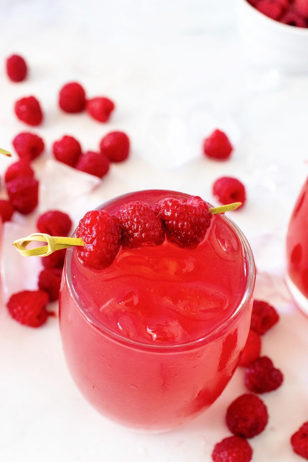 Slightly angled overhead shot of a glass filled to the brim with raspberry iced tea, garnished with a skewer holding three fresh raspberries. Scattered fresh raspberries and ice cubes surround the glass on a white and gray marble surface, with part of another glass and a bowl of fresh raspberries just visible off to the side.