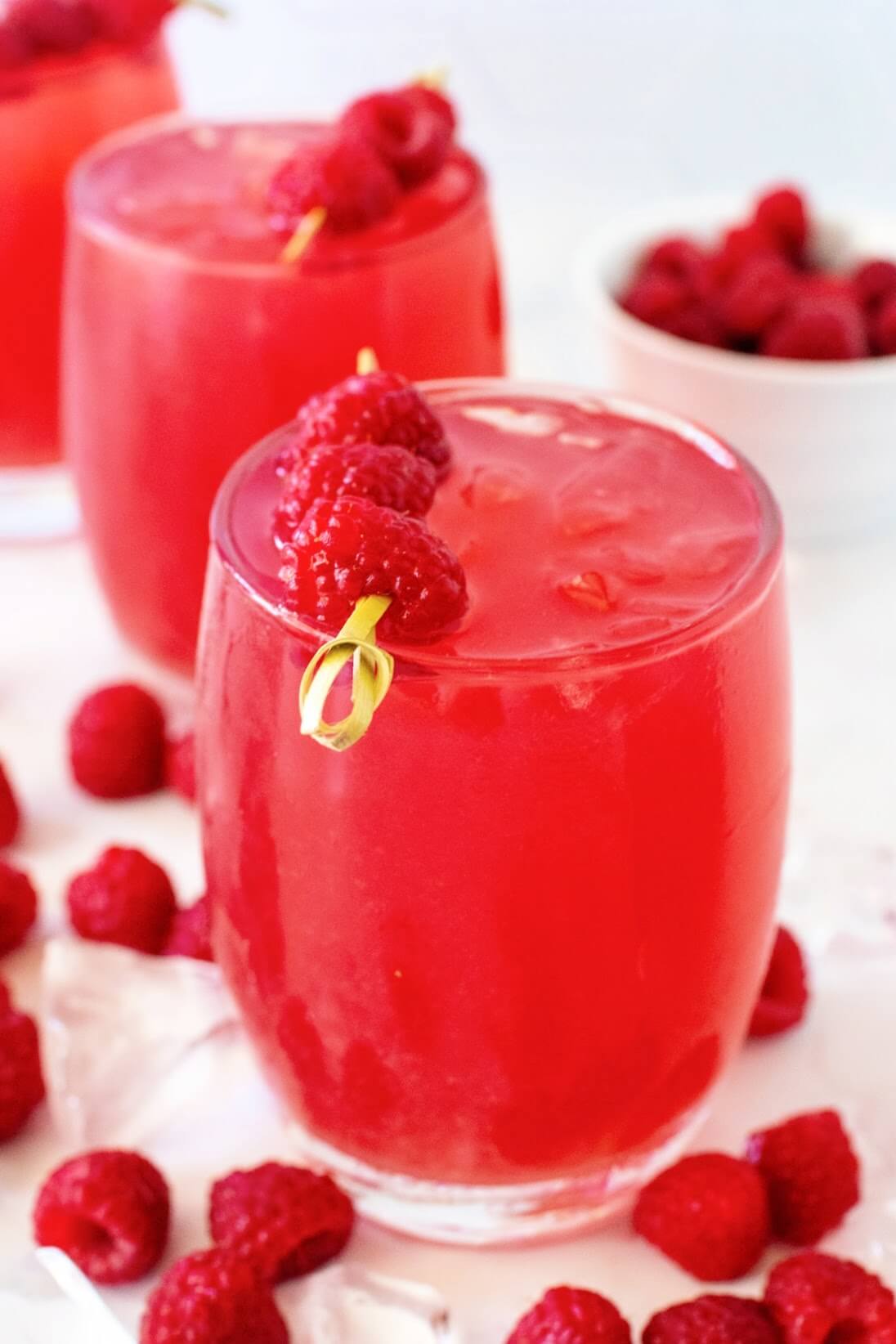 Slightly angled front view of a glass filled to the brim with raspberry iced tea, garnished with a skewer of three fresh raspberries. Fresh raspberries and ice cubes are scattered around it, with two more garnished glasses of raspberry iced tea and a white bowl of raspberries in the background, all placed on a white and gray marble surface.