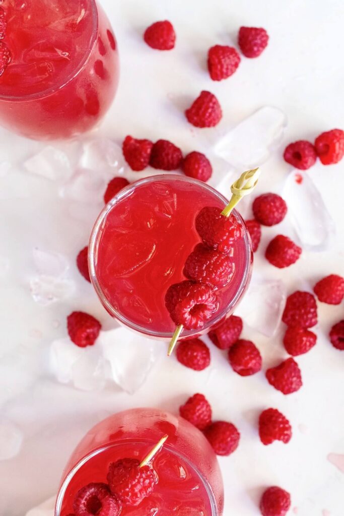 Close-up overhead shot of three glasses filled with raspberry iced tea, with two glasses partially off to the side. Two of the glasses are garnished with skewers holding three fresh raspberries. Fresh raspberries and ice cubes are scattered around them on a white and gray marble countertop.