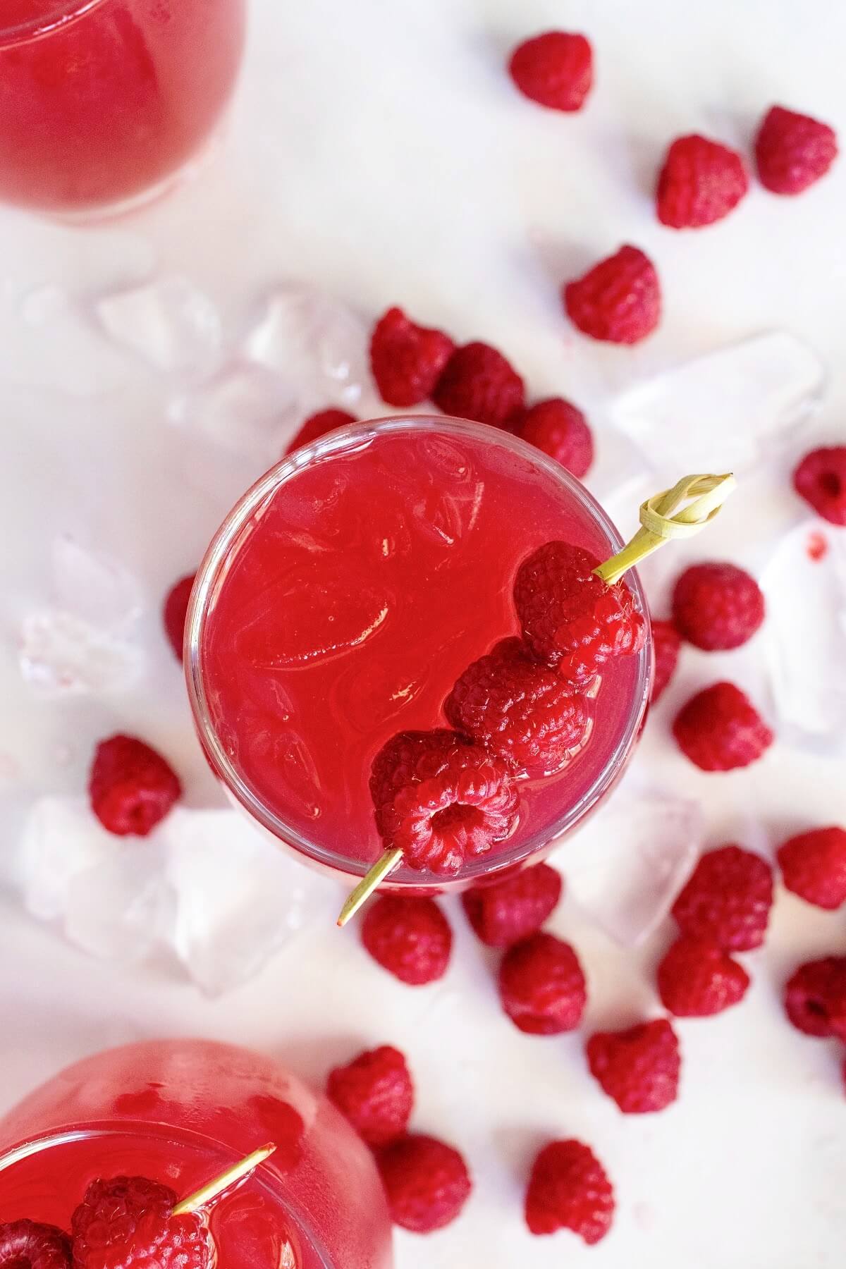 Close-up overhead shot of three glasses filled with raspberry iced tea, with two glasses partially off to the side. Two of the glasses are garnished with skewers holding three fresh raspberries. Fresh raspberries and ice cubes are scattered around them on a white and gray marble countertop.