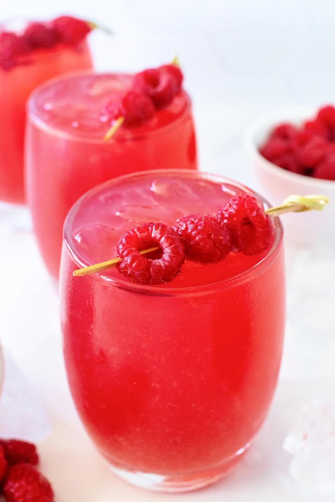 Front angled closeup view of a glass filled with raspberry iced tea, garnished with a skewer holding three fresh raspberries. A few fresh raspberries and ice cubes are scattered around it, with two more garnished glasses of raspberry iced tea and a white bowl of raspberries in the background, all set on a white marble surface.