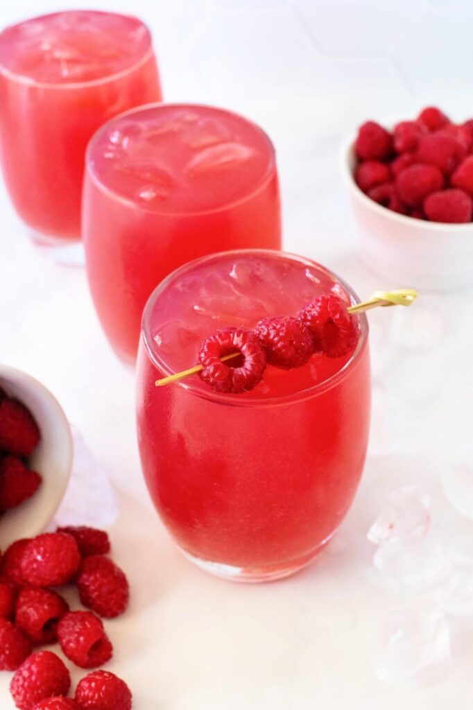 Front angled view of a glass filled with raspberry iced tea, garnished with a skewer holding three fresh raspberries. Ice cubes are scattered around it as well as a white bowl tipped over with fresh raspberries spilling out. In the background, two glasses of raspberry iced tea without garnishes sit near a white bowl filled with fresh raspberries — all on a white marble surface.