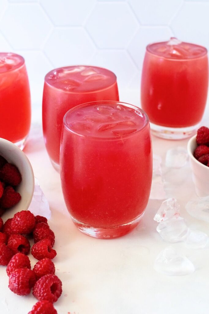 A white bowl tipped over in the front with fresh raspberries spilling out, next to a glass of raspberry iced tea filled to the brim with scattered ice around it. A partially visible bowl of raspberries is off to the side, with three more glasses of raspberry iced tea in the background, all set on a marble countertop in front of a white tile wall.