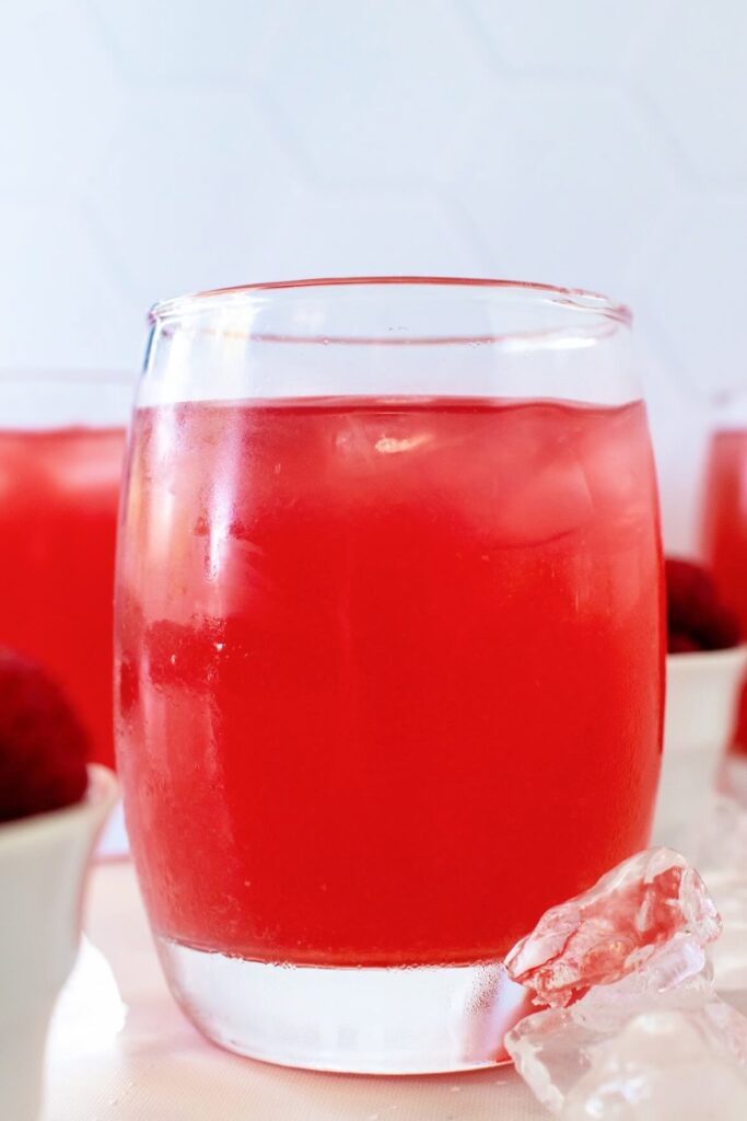 Close-up front shot of a glass filled with raspberry iced tea, with ice cubes scattered around it. Two white bowls filled with fresh raspberries sit nearby—one to the side and one behind the glass. In the background, two more raspberry iced tea glasses are blurred, with a white tile wall behind them, all on a marble countertop.