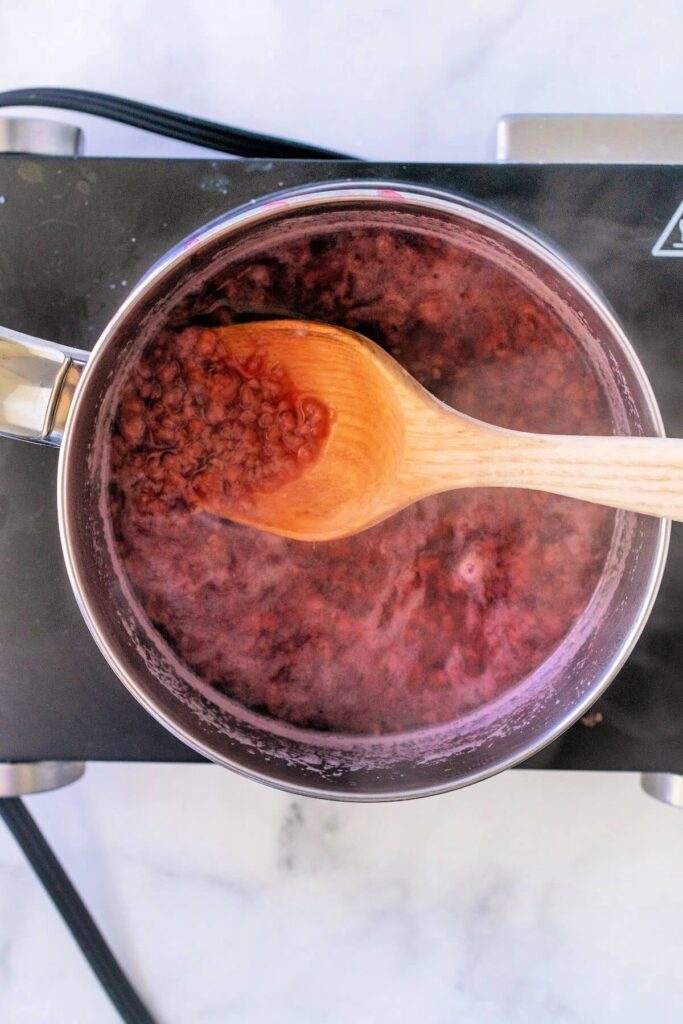 Stainless steel pan filled with a steaming hot, fully cooked and mashed raspberry mixture, sitting on a countertop electric burner on a white and gray marble surface.