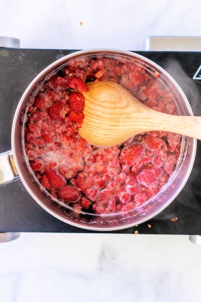 Stainless steel pan filled with a steaming hot mashed fresh raspberry mixture, sitting on a countertop electric burner placed on a white and gray marble surface.