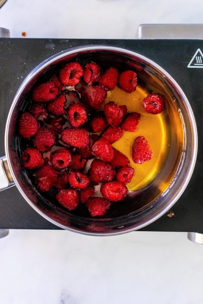 Stainless steel pan filled with fresh raspberries sitting in a honey-water mixture that resembles tea, placed on a countertop electric burner on a white and gray marble surface.