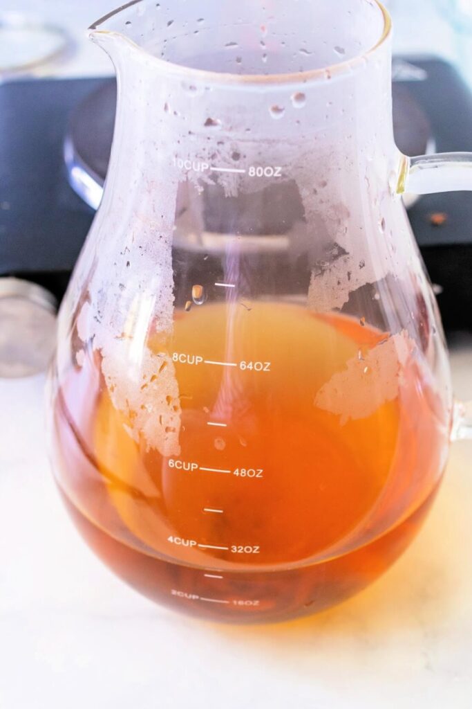 Glass pitcher filled with nearly 4 cups of steaming hot red raspberry leaf tea, placed in front of a countertop electric burner on a white and gray marble surface.
