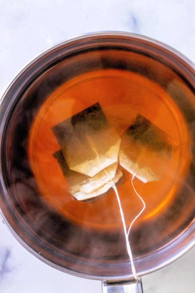 Overhead view of a stainless steel pan filled with fully steeped, steaming hot tea, with four red raspberry leaf teabags hanging in the pan. Set on a white and gray marble surface.