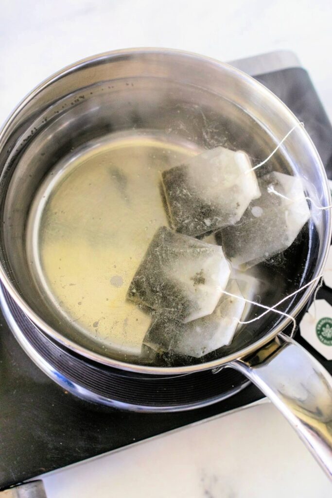 Overhead view of a stainless steel pan with steaming hot water and four red raspberry leaf teabags steeping, resting on a countertop electric burner over a white and gray marble surface.