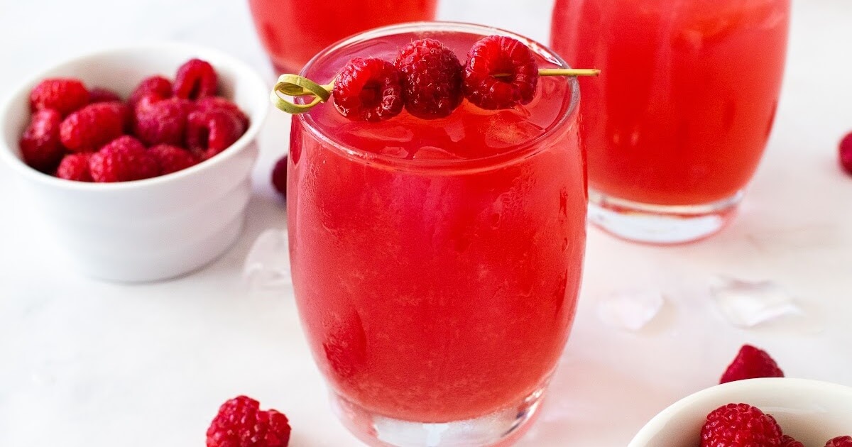 Closeup shot of a glass filled to the brim with iced raspberry tea, garnished with a skewer holding three fresh raspberries with a bowl of fresh raspberries just visible off to the front side. In the background, two more glasses filled with iced raspberry tea, a white bowl of fresh raspberries, scattered fresh raspberries, and ice cubes are arranged on a white and gray marble countertop.